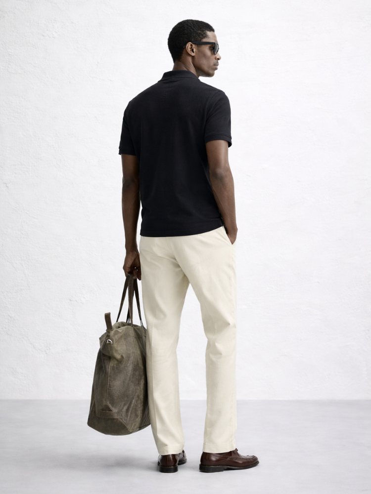 Man wearing a black polo shirt and white pants, holding a brown bag on a white background