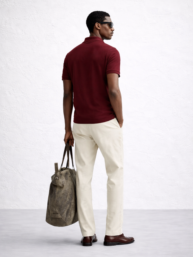 Man wearing a maroon polo shirt and white pants, holding a gray bag on a light background