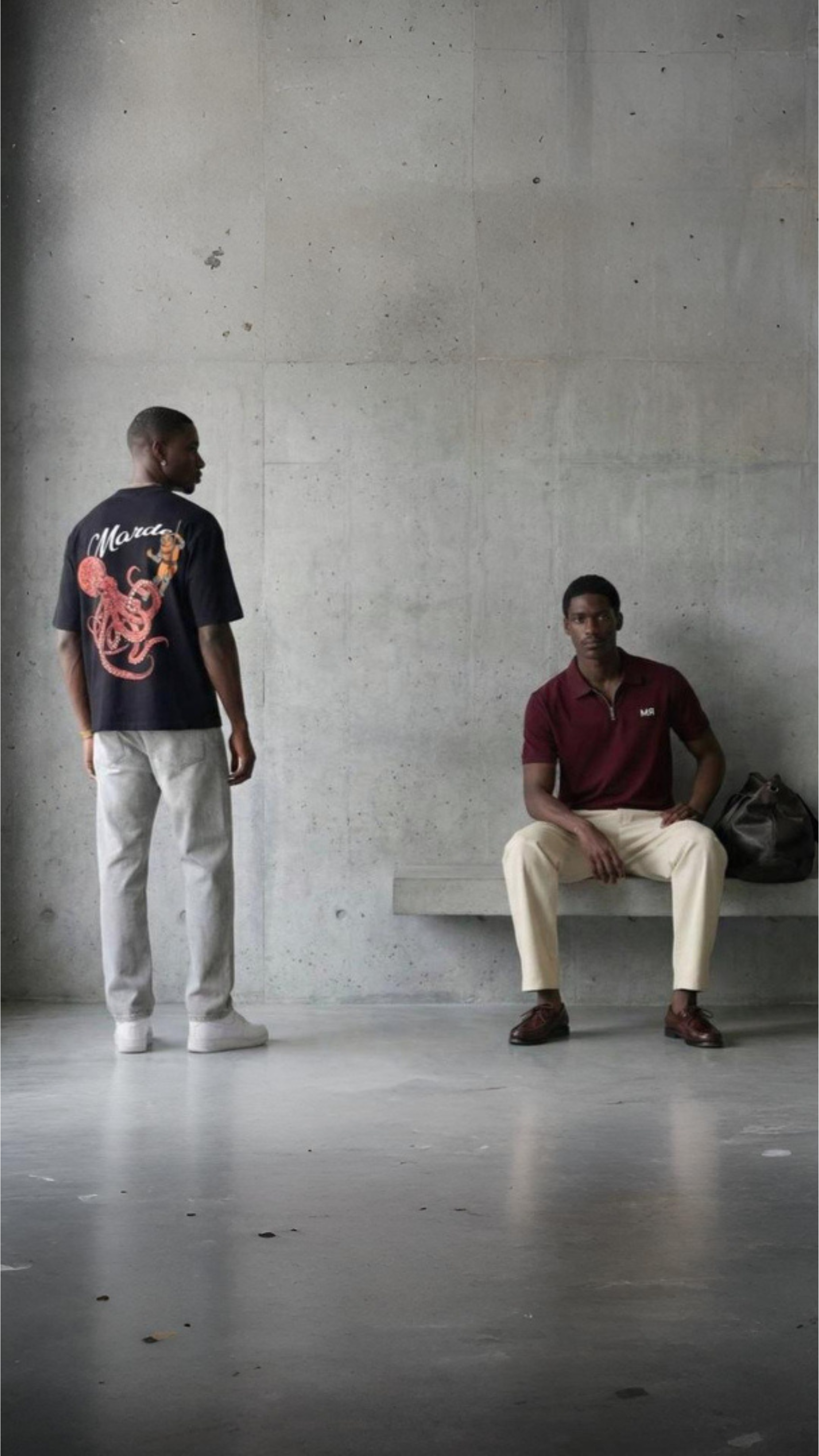 Two men in casual clothing standing and sitting against a concrete wall.
